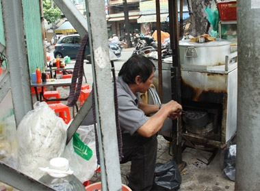 Ông Lương Tấn Túc (53 tuổi, quê Quảng Ngãi) bỏ than vào lò đốt để giữ nóng cho nồi nước lèo, phía sau là dãy bàn ghế vắng khách - Ảnh: Hữu Khoa